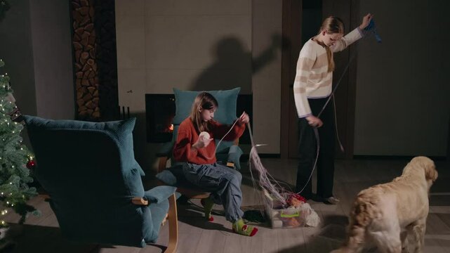 Two teenage girls untangling yarn with their family dog nearby, enjoying a cozy winter evening in a warmly lit room with a Christmas tree and fireplace.