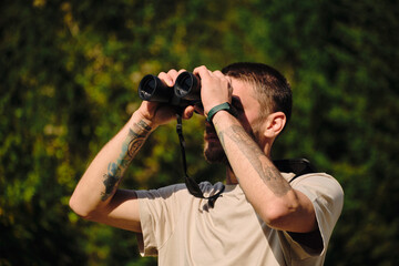 Young hiker man using binoculars while standing on a lookout in autumn forest. Bearded traveler guy holding binoculars outdoor. A man watches birds and wildlife.