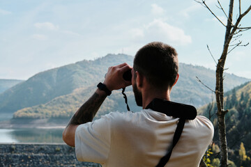 Obraz premium Young hiker man using binoculars while standing on a lookout in autumn mountains. Bearded traveler guy outdoor. A man watches birds and wildlife. Rear view. Uvac canyon in Serbia country.