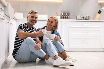 Obraz premium Happy middle aged couple with cups of drink on floor in kitchen, space for text