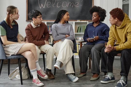 Full length front view portrait of multiethnic group of children with female therapist sitting on chairs in circle during group therapy session in school