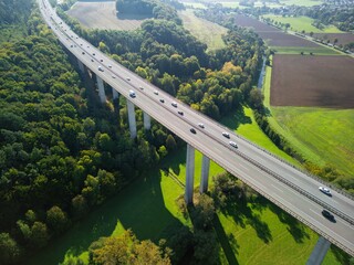 aerial view of a road bridge in Germany, running through a picturesque valley. The bridge is supported by tall concrete pillars that span green fields and forest. Rural settlements and small towns are