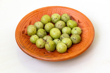 Amla fruits (Phyllanthus emblica), also called Indian gooseberry, are displayed in a clay plate on a light wood table.