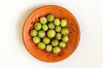 A clay plate containing Phyllanthus emblica (amla), also known as Indian gooseberry, emblic, emblic myrobalan, or myrobalan, sits atop a light wooden surface. Top view.