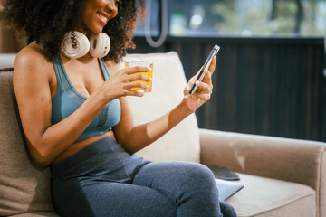 An African American woman in sportswear sits on a sofa near the dining table, preparing a healthy salad with vegetables fruits, drinking juice after exercise for weight control and low-fat nutrition