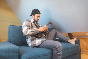 Obraz premium Man with talent sits on a gray sofa, carefully tuning a black ukulele. Musician is enjoying the feeling of playing the ukulele and immersing oneself in the creative spirit