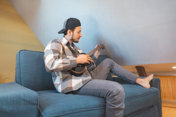 Obraz premium Man with talent sits on a gray sofa, carefully tuning a black ukulele. Musician is enjoying the feeling of playing the ukulele and immersing oneself in the creative spirit