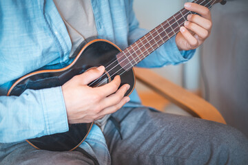 Fototapeta premium Close-up perspective captures a man's fingers plucking the strings of a glossy black ukulele. Making a music. Pleasure of playing an instrument