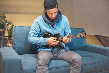 Musician wearing a beanie and denim jacket sits on the floor, strumming a ukulele with focus and...