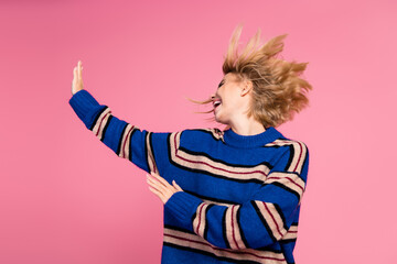 Joyful woman with blond hair laughing in a bright striped sweater amid a pink background expressing happiness and motion