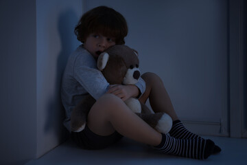 Fearful boy hiding with teddy bear on windowsill at night