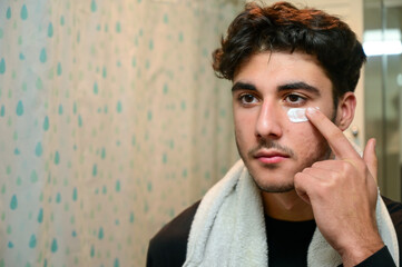 Man applying face skincare cream while grooming looking at the mirror in the bathroom in his morning routine at home. Close up portrait of handsome guy using facial lotion for clean and clear skin