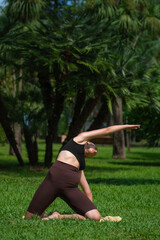 Young woman in sports clothes does yoga in a green park, demonstrating an inclined stretch with her arm outstretched back on a yoga mat surrounded by nature. Concept of smart fitness, yoga, pilates