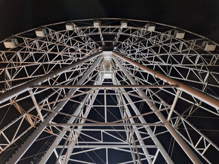 A view from below of a Ferris wheel against a dark sky. The metal elements of the structure are illuminated by a spotlight. Background.