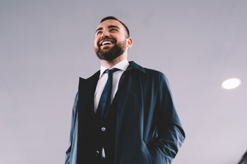 Below view of cheerful male entrepreneur 30 years old looking away and laughing indoors, successful Caucasian businessman dressed in formal clothing and trendy coat have work trip from airport