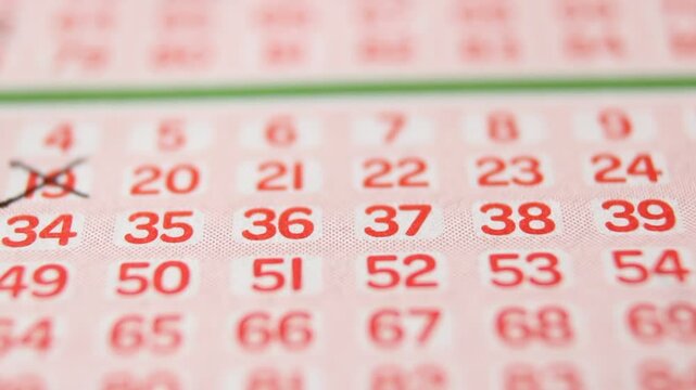 A man marks with a pen numbers 19, 36, 66, 39, 68 on a lottery ticket. The rows of red numbers on the white lottery ticket are arranged horizontally. Close-up horizontal video