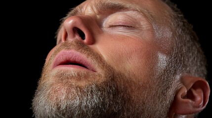 Obraz premium Close Up of Man Exhaling: A close up shot of a man's face as he exhales, his eyes closed and his beard visible against a dark background.