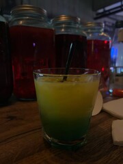 A vibrant cocktail sits on a rustic wooden table, featuring a colorful gradient from yellow to green. Various jars in the background set a lively atmosphere for the drink.