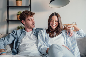 Young happy couple having fun talking laughing relaxing at home on couch, boyfriend embracing...