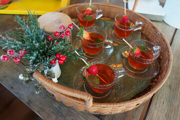 Five glass cups filled with herbal drinks in a woven tray. Each cup is adorned with a strawberry and mint, set amidst charming decorations.