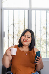 portrait of plus size mexican woman at home holding coffee cup and mobile phone, overweight model