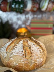 A specialty holiday loaf of sourdough, with skillfully scored Christmas trees around the golden brown crust of the bread. A candle glows in the background adding to the cozy winter feel.