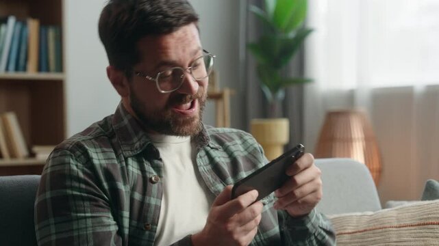 Caucasian adult gamer gadget addict man male guy playing virtual video game play mobile phone gaming excited having fun online cyber-sport championship at home couch relaxing smartphone entertainment