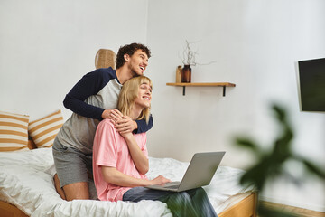 Two men share a loving embrace while using a laptop in a comfortable bedroom setting.