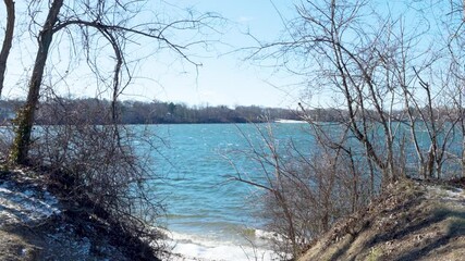 vA peaceful winter scene at Lake Ronkonkoma, Long Island. Clear blue skies reflect over shimmering icy waters, creating a tranquil moment of natural beauty. Captures the serene charm of the season.