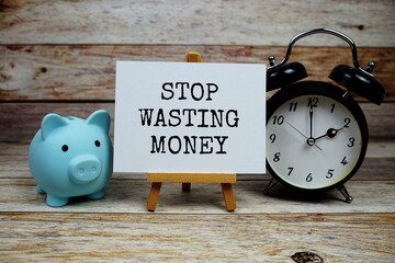 Stop Wasting Money text on paper card with alarm clock and piggy saving on wooden baciground, Money...