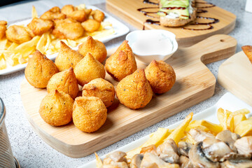 Coxinha, savory teardrop-shaped fritters filled with shredded chicken, displayed on wooden board alongside dipping sauce