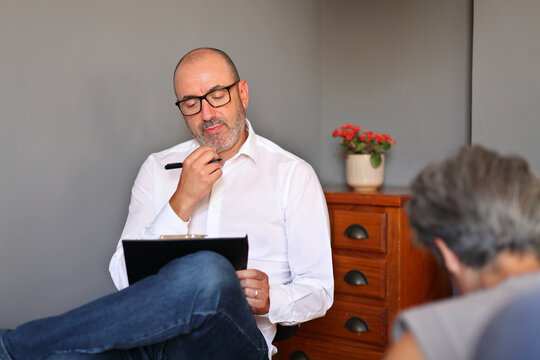 A therapist listens attentively and takes notes during a confidential meeting with a mature female client in a modern office.