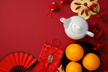 Chinese New Year celebration items on red background: white teapot, oranges, red fan, red envelope with coins, lanterns, and cherry blossoms