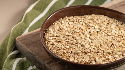 Brown ceramic bowl filled with rolled oats placed on a rustic wooden board, with a green striped kitchen towel in the background. Healthy breakfast concept.