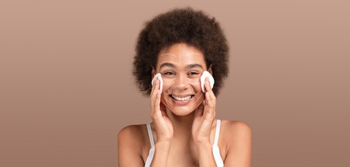 Fototapeta premium African American woman enjoys her skincare routine, applying cleanser on her face with cotton pads. She is isolated against a solid background, radiating joy during her pampering session.