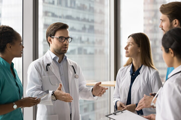 Young male general practitioner in coat standing in clinic with staff members, explain procedure,...