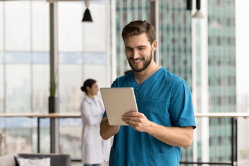 Young man medical worker wear blue scrubs using tablet in clinic, leading telemedicine consultations, conducting virtual meetings with patients, prescribing medications remotely, managing appointments