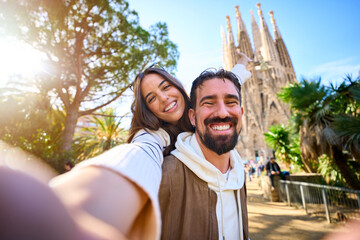 Young mixed race couple taking a selfie in front of Sagrada Familia in Barcelona. Smiling Husband and wife on street of city enjoying vacation happy and smiling. Funny tourism people