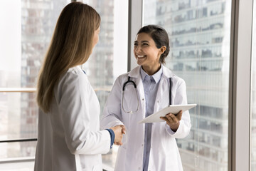 Two happy multiethnic women therapists, clinic colleagues standing in modern skyscraper office...
