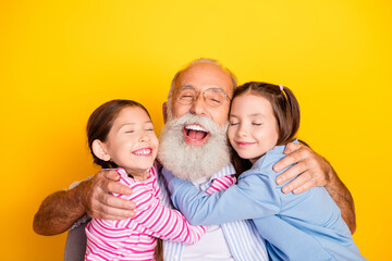 Grandfather joyfully hugging two young granddaughters on a bright yellow background with happy, loving expressions.
