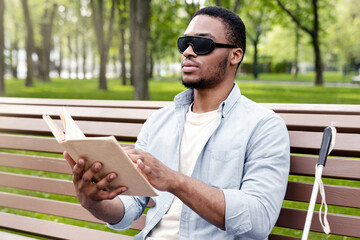 Young black visually impaired man sitting on bench in city park, reading Braille book outdoors....