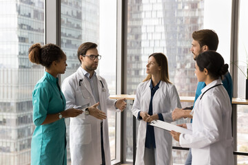 Serious staff members, clinic workers dressed in uniforms stand together in office, listen speech...