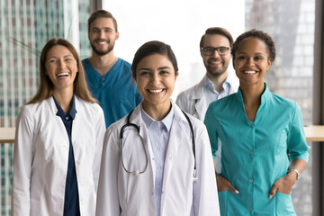 Young Indian female doctor dressed in white lab uniform stands at front, smiling confidently, with her diverse colleagues in background. Leadership, mission, team spirit. Healthcare workers portrait