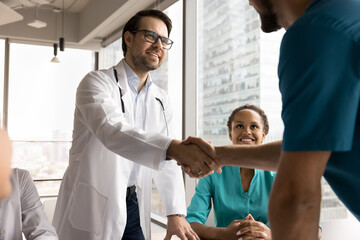 Obraz premium Two male doctors in uniforms shaking hands, start medical concilium in clinic office boardroom with diverse staff members. Gesture of mutual respect, agreement, or conclusion of successful discussion