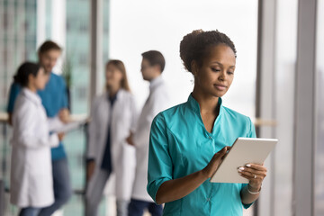 Focused African doctor dressed in scrubs uniform using tablet, accessing records, documenting patient visits, symptoms, and treatments plans, viewing health test results. Medicine, modern, tech usage