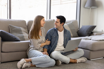 Cheerful young girlfriend and boyfriend enjoying leisure together, holding laptop computer, sitting on floor at couch, using smart home application, wireless Internet technology, talking, laughing