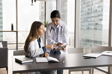 Two women, Caucasian and Indian female GP professionals wear white lab coats working together using...