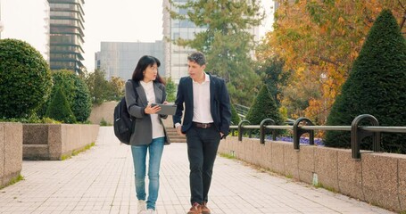 Two professionals, man and woman, engage in discussion while strolling through a modern urban park in the financial district, sharing insights in a tablet while enjoying a productive atmosphere