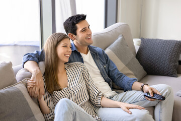 Obraz premium Positive attractive young couple watching romantic movie on TV at home together, resting on comfortable sofa, hugging, talking, laughing, using remote control