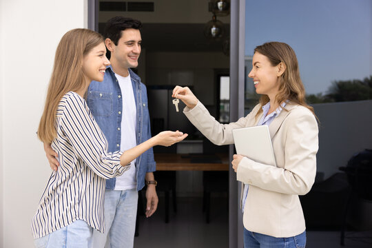 Confident female professional realtor woman giving key to couple of customers, offering rental to happy young husband and wife, meeting with new property owners at doorstep